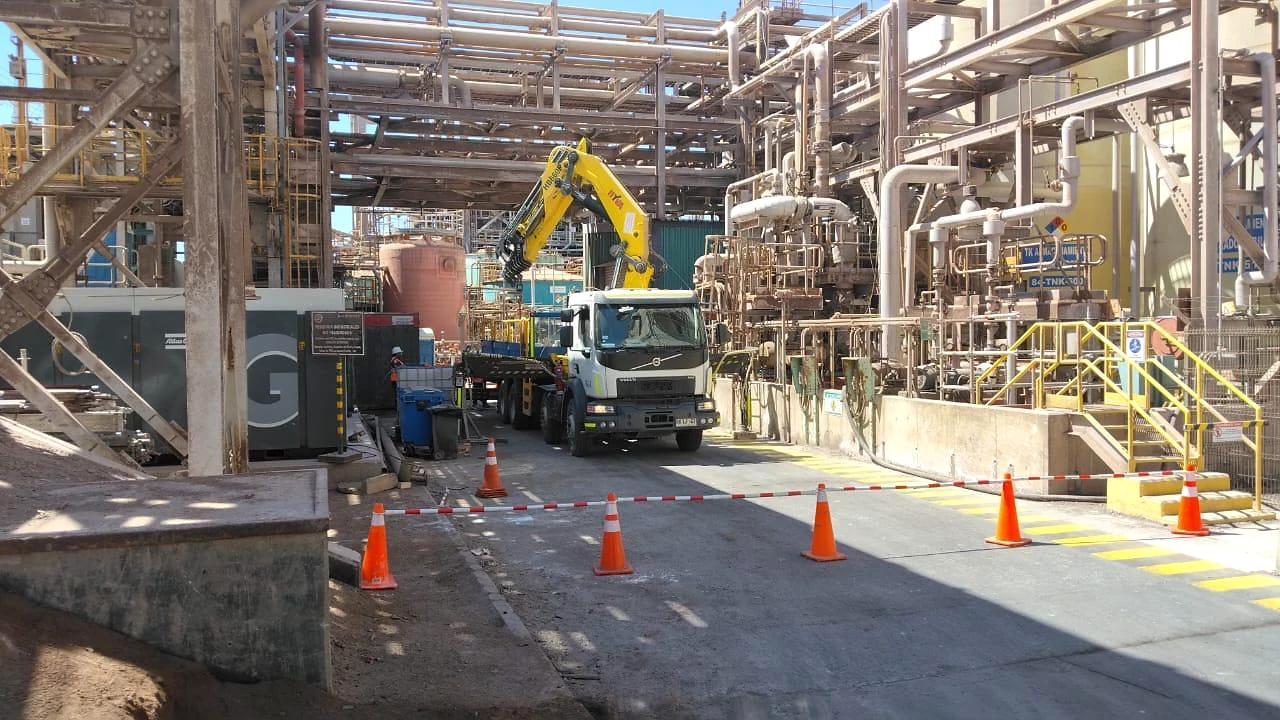 Operación en instalación industrial. Camión pluma ejecutando apoyo de izaje entre estructuras de proceso.